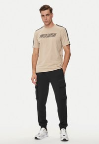 Maglietta in cotone beige con logo nero "GUESS", abbinata a pantaloni cargo neri con tasche laterali, indossata con scarpe sportive bianche.