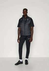 Czarna kamizelka izolacyjna z detalami w diamentowy wzór, noszona na czarnej koszulce z krótkim rękawem i czarnych spodniach dresowych, połączona z czarnymi butami sportowymi.