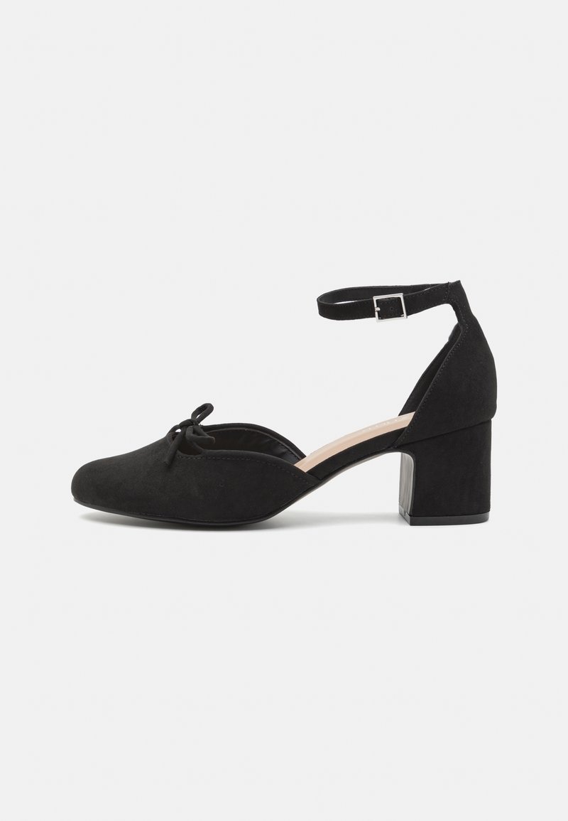Chaussure à talon bloc en daim noir à bout fermé avec bride à la cheville et petit nœud décoratif sur le dessus, vue de côté sur fond blanc.
