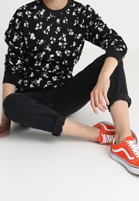 Černá mikina s květinovým vzorem a dlouhými rukávy, spojená s ohrnutými černými kalhotami a oranžovými teniskami. Hladký povrch; neutrální pozadí.