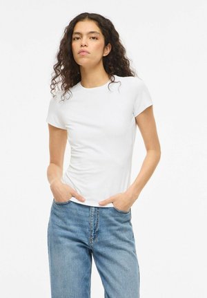Jeune femme aux cheveux bouclés portant un t-shirt blanc uni et un jean bleu, debout les mains dans les poches sur un fond blanc.