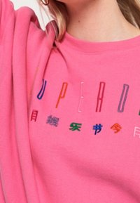 Pink sweatshirt featuring multicolored embroidered text on the chest, with a crew neck and relaxed fit. Soft fabric texture.