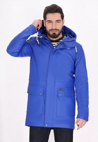 Niebieska wodoodporna kurtka z kapturem, zapinana na zamek z przodu, z dwiema dużymi kieszeniami z przodu i akcentami w formie sznurka. Noszona na w paski koszulką.