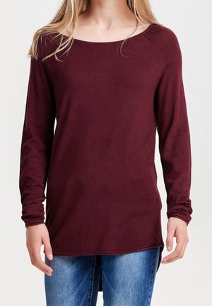 Vrouw die een langarmige, bordeaux tuniektrui draagt in combinatie met een blauwe spijkerbroek, staand tegen een effen lichte achtergrond.