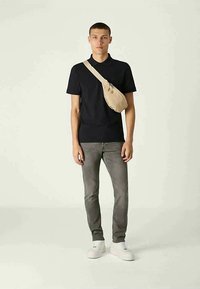 Marineblaues Poloshirt, graue Jeans, weiße Sneakers und eine beigen Umhängetasche, die über der Schulter getragen wird, vor einem neutralen Hintergrund stehend.