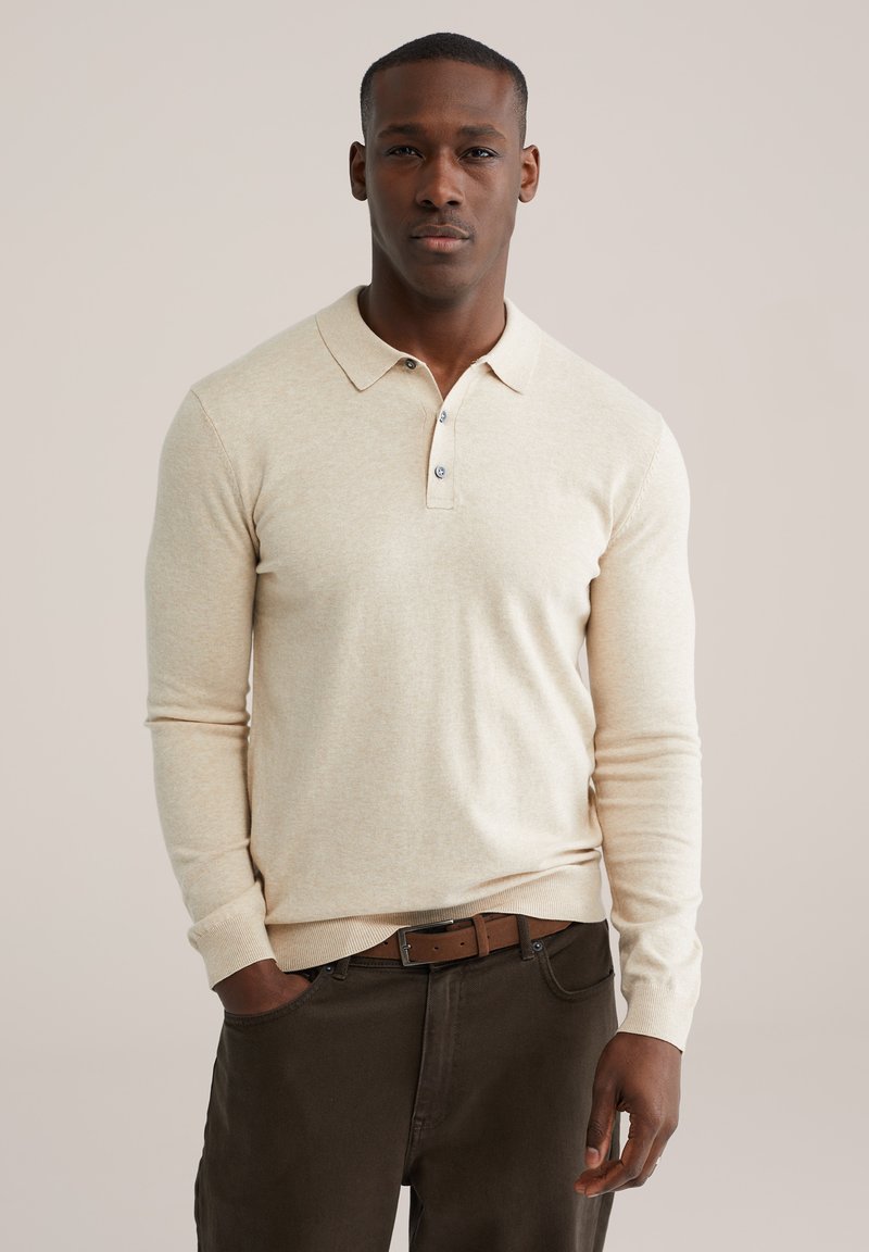 Mann trägt ein beigefarbenes langärmeliges Poloshirt, das in eine dunkelbraune Hose mit braunem Gürtel gesteckt ist, und steht mit einer Hand in der Hosentasche.