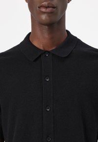 Black polo shirt with a textured fabric, featuring a collar and six buttons down the front, displaying a sleek, minimalist design.