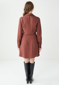Brązowa sukienka z długim rękawem, dopasowaną talią, kołnierzykiem oraz zwiewnym dołem. Noszona z czarnymi kozakami do kolan. Gładka tekstura materiału.