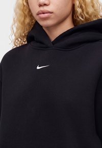 Човек с къдраво русо коса, носещ черен суичър с малко бяло лого на Nike на гърдите.