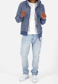 Mężczyzna w białych sneakersach, jasnoniebieskich dżinsach, białej koszulce i niebieskiej bluzie z zamkiem, trzymający sznurki od bluzy.