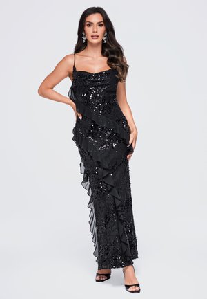 Femme portant une robe noire sans manches à sequins avec des détails diagonaux à volants, debout avec une main sur la hanche et portant des boucles d’oreilles remarquables.