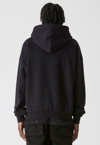 Fekete kapucnis pulóver laza szabással, bordázott mandzsettával és szegéllyel, valamint textúrált anyagból készült. A dizájn jellemzője egy nagy hátsó kapucni.