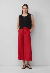 Gilet noir au crochet avec bouton à l'avant, associé à un pantalon rouge ample et large. Le modèle porte des sandales noires plates. Fond uni.