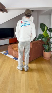 Λευκή μακριά μπλούζα με μπλε και κόκκινο λογότυπο "Cortina Dolomiti". Ανοιχτό denim παντελόνι, casual αθλητικά παπούτσια, εσωτερικό περιβάλλον με φυτά.