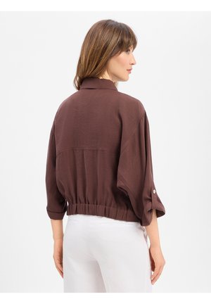 Vrouw draagt een losse bruine jas met knopen op de mouwen en een elastische taille, witte broek, staat iets gedraaid naar de zijkant tegen een effen achtergrond.