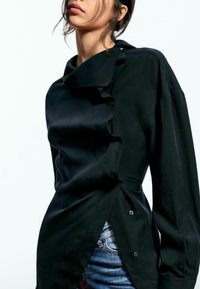 Bluză neagră cu guler înalt, cu detalii volănașe și închidere cu butoane pe lateral. Fabricată dintr-un material neted, cu mâneci lungi.