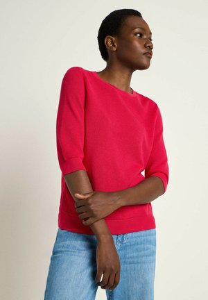 Bright pink knit sweater with a round neckline, three-quarter sleeves, and a textured pattern. Paired with light blue denim.