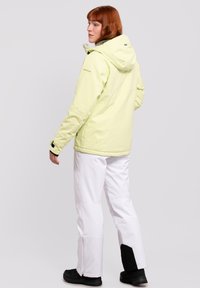 Hellgrüne, wasserdichte Jacke mit Kapuze, elastischen Bündchen und Reißverschlusstaschen, kombiniert mit weißen Hosen, die schwarze Akzente im unteren Bereich aufweisen.