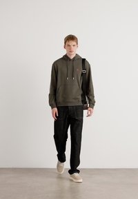 Felpa verde oliva con cordini regolabili, tasca frontale; abbinata a pantaloni neri e sneakers beige; modello che tiene in mano una borsa nera.