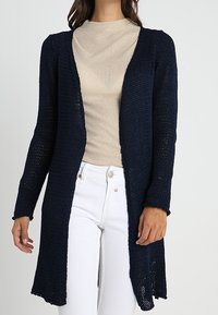 Marinblå stickad cardigan med öppen framsida, långa ärmar och sidoflikar, bärs över en beige topp och vita byxor, med en lös vävstruktur.