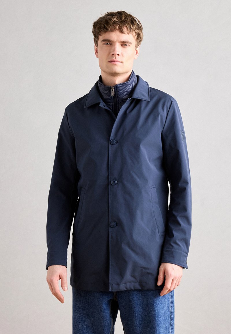 Cappotto impermeabile blu navy con chiusura a bottoni, ampio colletto, tasche laterali e foderatura interna a contrasto, indossato sopra una giacca con zip.