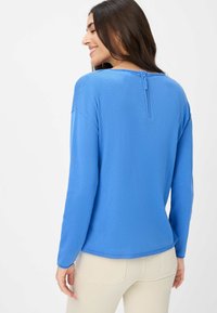 Blauwe lange mouw top met een rits aan de achterkant, zachte textuur en een iets ontspannen pasvorm. Eenvoudig ontwerp; geen zichtbare patronen of accenten.