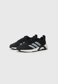 Chaussures de sport noires avec un dessus en mesh texturé, dotées de rayures grises, d'un col rembourré et d'une semelle en caoutchouc blanche avec une semelle extérieure noire.