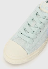 Gros plan d'une sneaker Converse All Star bleu clair montrant les lacets, le tissu en daim et les détails en caoutchouc blanc sur la pointe et la semelle.