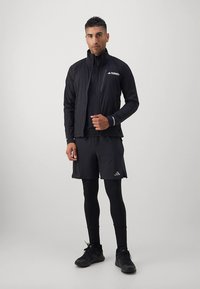 Casaco atlético preto com tecido texturizado, fecho de correr completo, gola alta, combinado com calções pretos e leggings, apresentando um logótipo no casaco e nos calções.