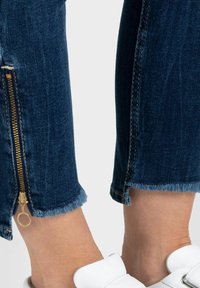 Jean en denim bleu foncé avec un ourlet effiloché, présentant un détail de fermeture éclair dorée à la cheville. Une chaussure blanche est partiellement visible en bas.