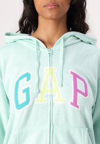 Mintgrønne lynlås hoodie lavet af blødt stof. Har flerfarvede broderede "GAP" bogstaver i turkis, gul og pink. Foran lommer inkluderet.