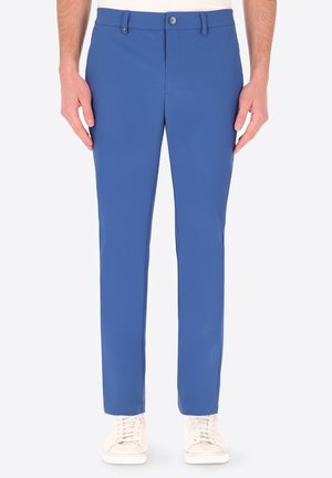 Pantaloni blu a gamba dritta indossati con sneakers bianchi, abbottonati in vita, mostrati da metà torso ai piedi su sfondo bianco.