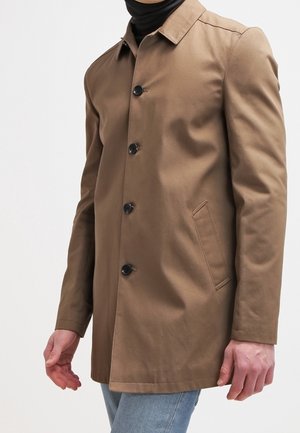 Homme portant un manteau marron boutonné sur un col roulé noir et un jean bleu clair sur un fond blanc uni.
