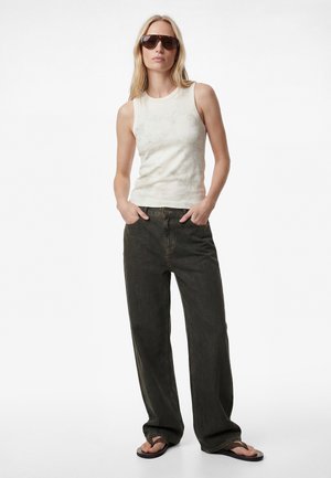 Kobieta ubrana w bezrękawnik w jasnobeżowym kolorze, ciemne szerokie jeansy, czarne klapki i duże brązowe okulary przeciwsłoneczne, stoi z rękami w kieszeniach.