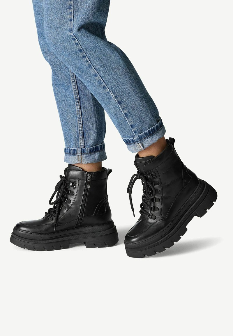 Bottines noires en cuir avec une semelle épaisse, un laçage à l'avant, une fermeture éclair sur le côté et un col rembourré. Portées avec un jean en denim bleu retroussé.