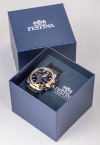 Auksinis ir sidabrinis chronografas laikrodis tamsiai mėlyname dėkle su logotipu. Laikrodis turi mėlyną ciferblatą, tekstūruotą dirželį ir datos rodymą.