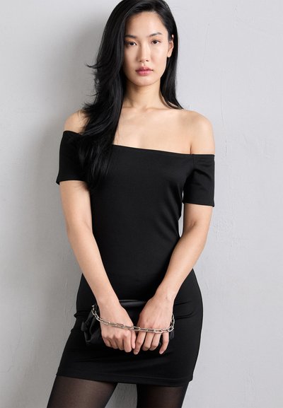 Vestido negro sin hombros con mangas cortas, silueta ajustada y textura suave. La modelo sostiene un clutch negro con un acento de cadena.