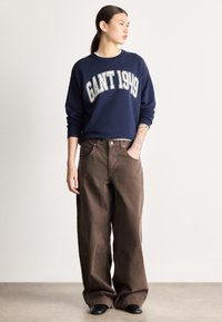 Tengerészkék pulóver fehér "GANT 1949" grafikával, laza barna nadrággal és fekete lábbelivel párosítva; csípőig érő fazon és kényelmes szabás.
