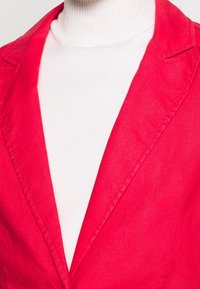 Red linen blazer with a notched collar, layered over a white ribbed turtleneck. The fabrics showcase a smooth texture and vibrant colors.