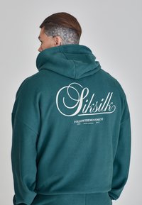 Türkiz kapucnis pulóver, hátul nagy fehér logóval, amelyen "Siksilk" írásjel formájában, alatta további szöveggel. Lágy, texturált anyag.