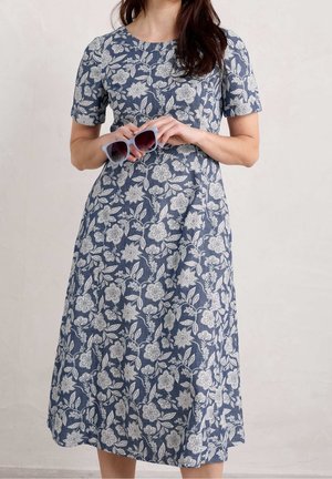 Vrouw die een blauwe midi-jurk met wit bloemmotief draagt en lichtblauwe zonnebril met paarse glazen vasthoudt.