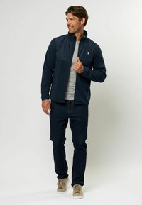 Giacca in pile blu navy con colletto alto, zip frontale e dettaglio sul pannello del petto, abbinata a jeans blu scuro e sneakers beige.
