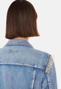 Chaqueta de mezclilla azul claro con un diseño estructurado, adornada con un acento de cadena y un detalle de flecos transparentes en el hombro.