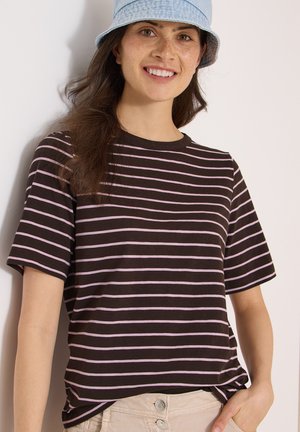 Jeune femme aux longs cheveux foncés, portant un chapeau bob bleu clair et un t-shirt rayé noir et rose, souriante avec une main dans la poche d'un pantalon beige.