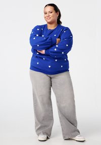 Sweat-shirt bleu roi avec motifs de cœurs blancs, associé à un jean large gris clair et des baskets blanches. Coupe confortable et décontractée.