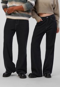 Jean large noirs avec des coutures contrastantes. Associés à un pull court crème et un pull tricoté multicolore. Bottes noires épaisses.