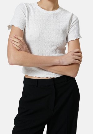 Persona che indossa una maglietta bianca a maniche corte con trama e pantaloni neri, con le braccia incrociate sul torso.