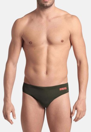 Uomo senza maglietta che indossa slip da bagno verde scuro con logo rosso "arena" sull'anca sinistra su sfondo chiaro e uniforme.