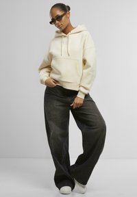 Krem hettegenser med foran lomme og justerbar snøring, sammen med oversized mørkegrå jeans og hvite sneakers. Tilbehør inkluderer solbriller.