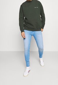 Sudadera verde oscuro con cuello redondo y texto blanco en el pecho, combinada con jeans ajustados azul claro y zapatillas blancas con tres rayas.